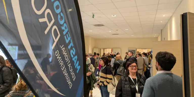 Una vela totem marchiata mastercom pro, a lato due persone che parlano in piano medio e sullo sfondo una moltitudine di persone all'interno di un'aula di una scuola