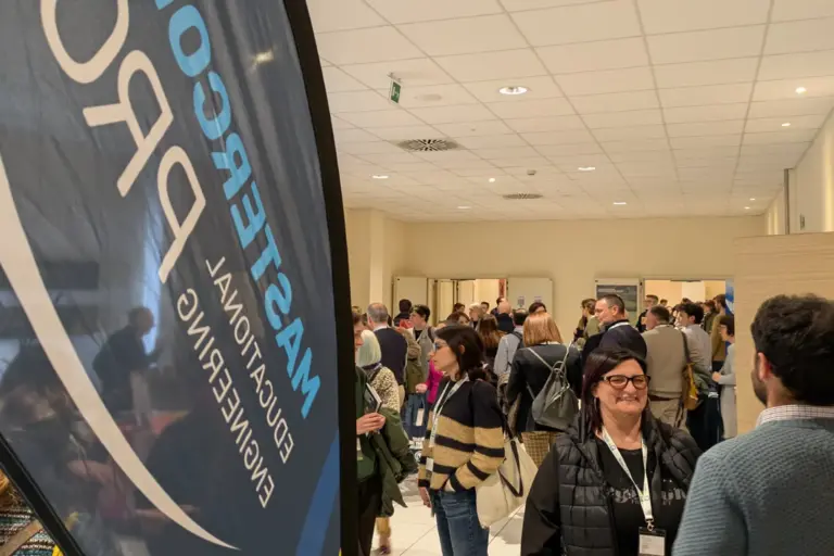 Una vela totem marchiata mastercom pro, a lato due persone che parlano in piano medio e sullo sfondo una moltitudine di persone all'interno di un'aula di una scuola