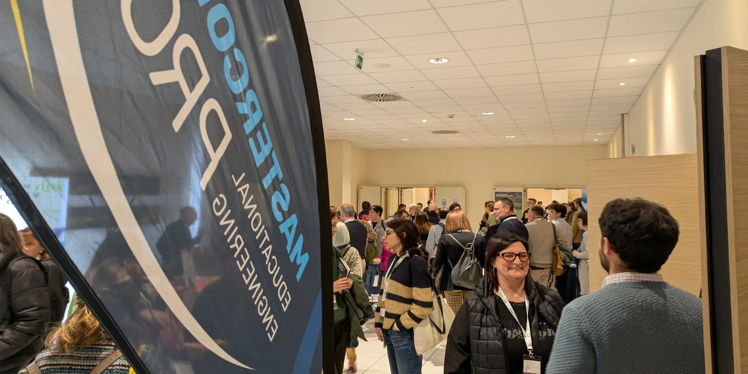 Convegno-Nazionale-FOE-2026–costruire-insieme-futuro-scuola Una vela totem marchiata mastercom pro, a lato due persone che parlano in piano medio e sullo sfondo una moltitudine di persone all'interno di un'aula di una scuola